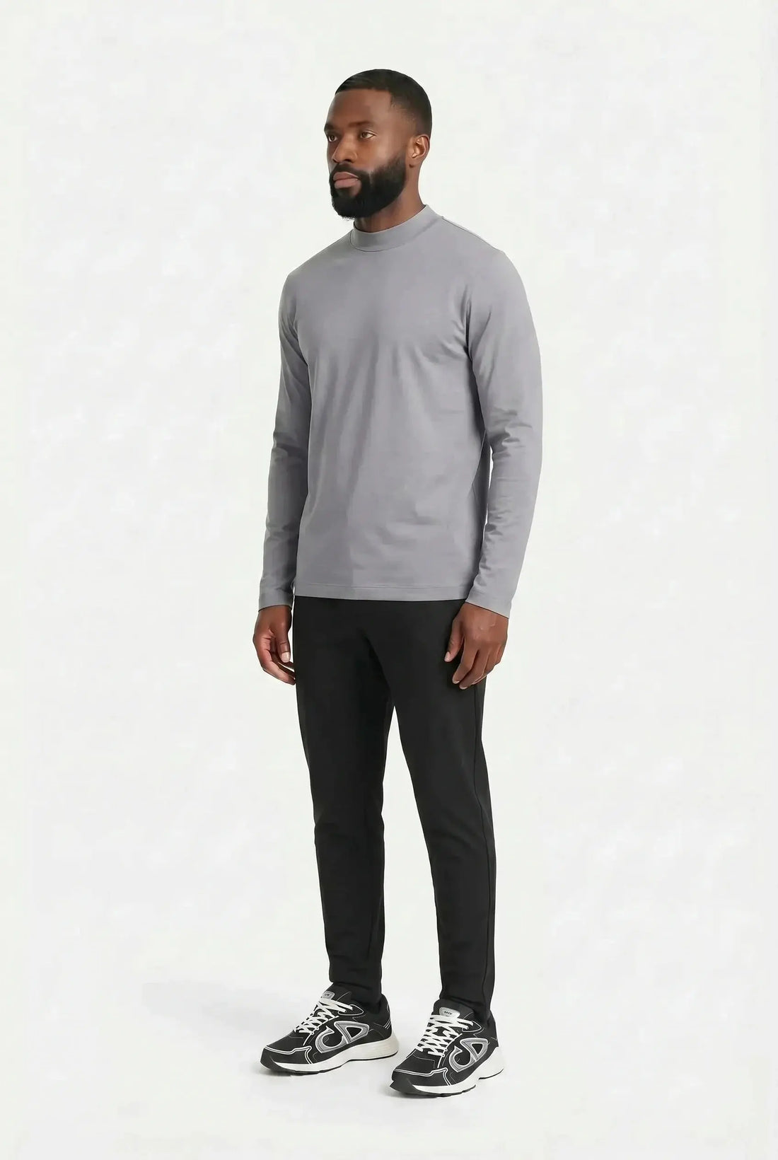 Homme debout de trois quarts, silhouette entière, en t-shirt à manches longues gris perle et pantalon noir, sur fond clair uni